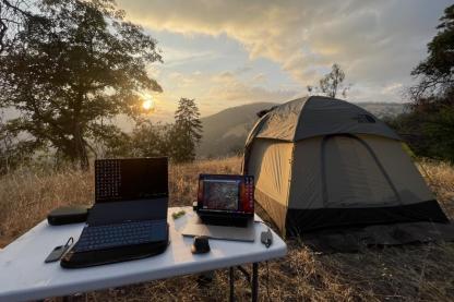 Laptop on a table outside next to a tent, with trees and hills in the background