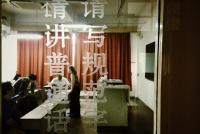 Looking through a window at a room with at people at desks and tables. On the window are Chinese characters that read please speak Mandarin, write proper characters